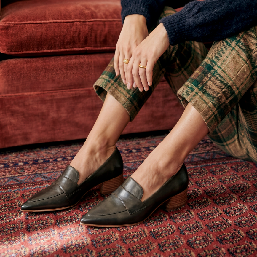 Person wearing Dark Olive loafers with a stacked leather heel on a patterned rug with a red couch in the background