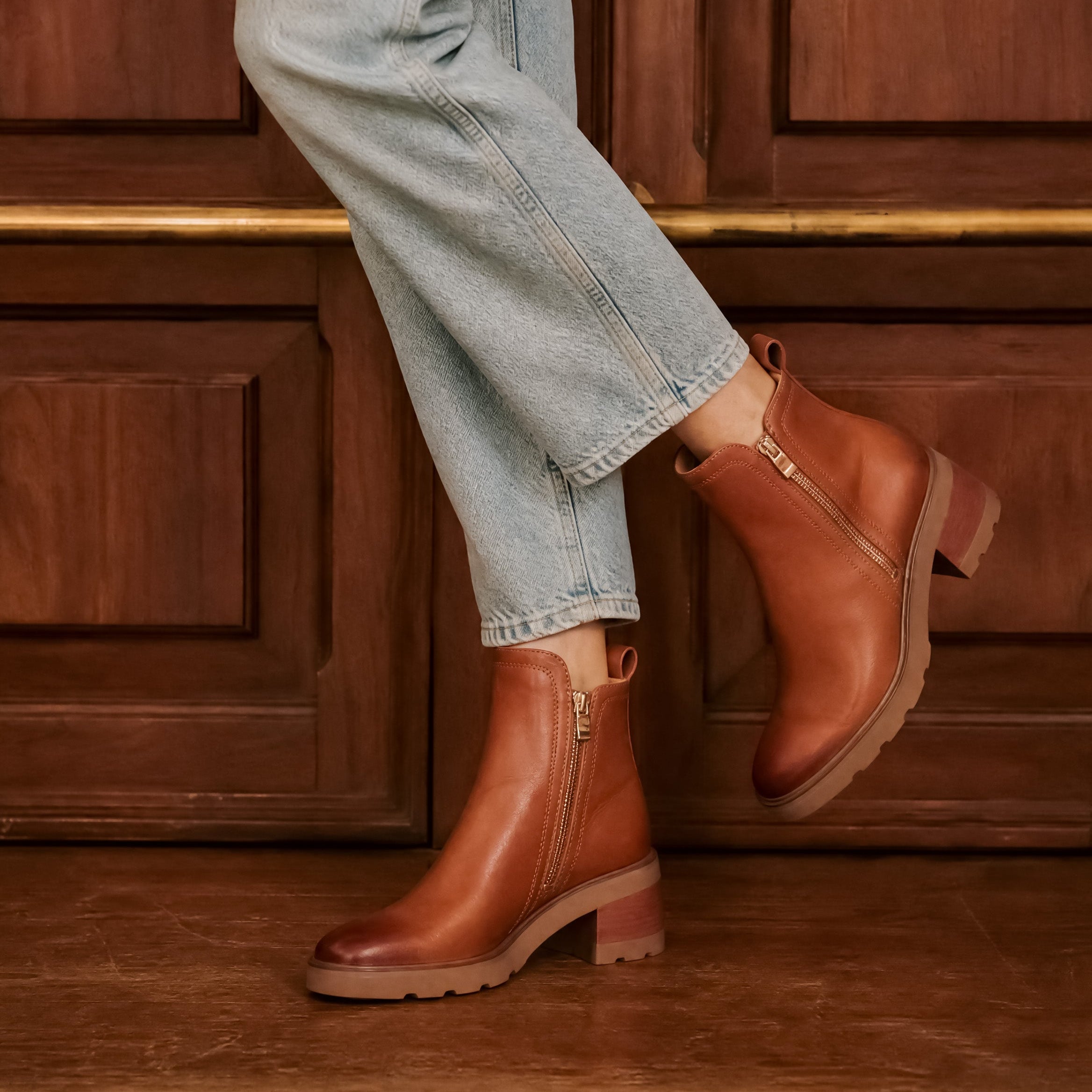 Person wearing brandy leather boots and light blue jeans against a wooden background
